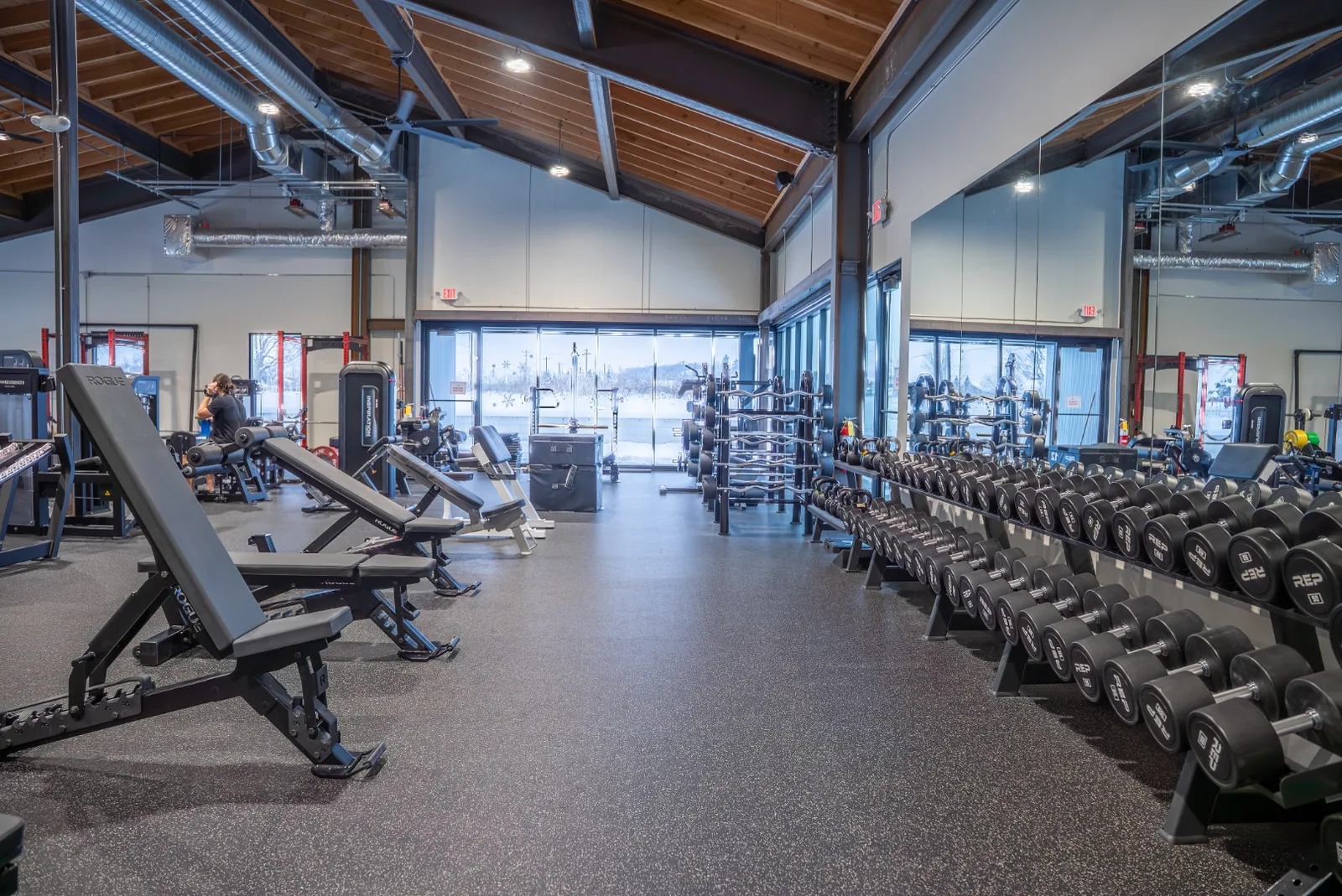 Rows of adjustable benches and dumbbell racks across the VirtueHall open-gym floor under the vaulted ceiling