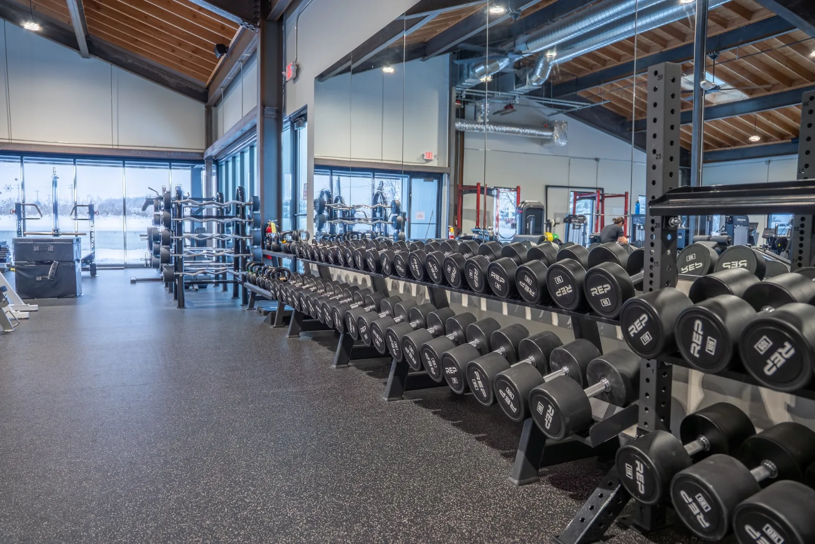 VirtueHall free-weight area with adjustable benches and a long dumbbell rack beside a wall of windows